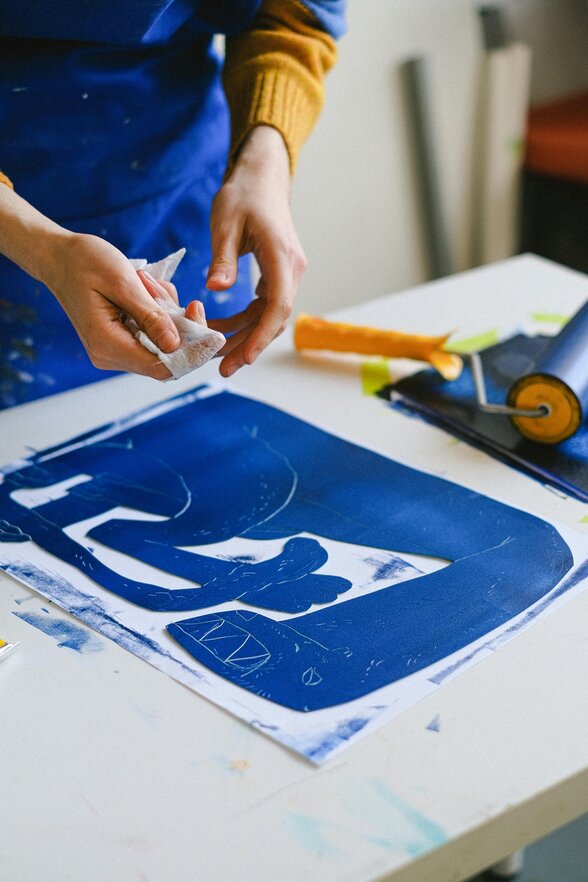 Mains manipulant un bloc encré en bleu sur un plan de travail, avec rouleau encreur et feuilles imprimées - Agrandir l'image, fenêtre modale