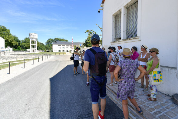 Un guide explique le site à un groupe de visiteurs réunis devant un bâtiment, par beau temps - Agrandir l'image, fenêtre modale