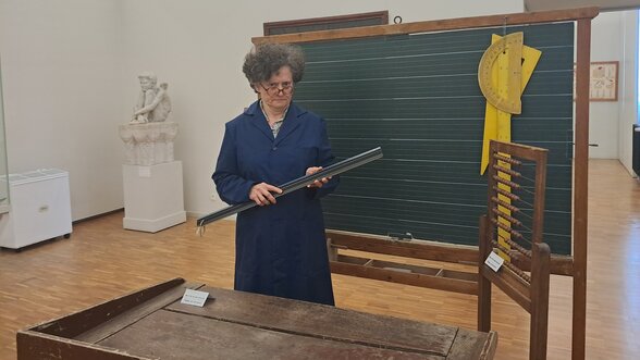 Une animatrice costumée en institutrice tient une grande règle devant un tableau noir, entourée de mobilier scolaire ancien - Agrandir l'image, fenêtre modale