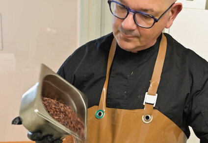 Artisan en tablier versant des éclats de fèves de cacao dans une cuve de broyage en inox - Agrandir l'image, fenêtre modale