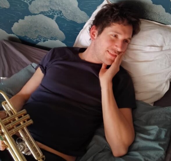 Marius Atherton, musicien, allongé et souriant, tenant une trompette dorée dans une pose décontractée - Agrandir l'image, fenêtre modale
