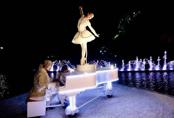 Danseuse en tutu blanc juchée sur un piano lumineux motorisé, fontaines illuminées en arrière-plan nocturne - Agrandir l'image, fenêtre modale