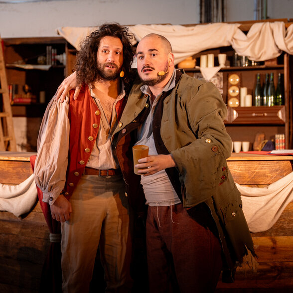 Deux comédiens en costumes d'époque posant complices sur un décor de taverne, micros HF visibles, lors d'une représentation en immersion. - Agrandir l'image, fenêtre modale