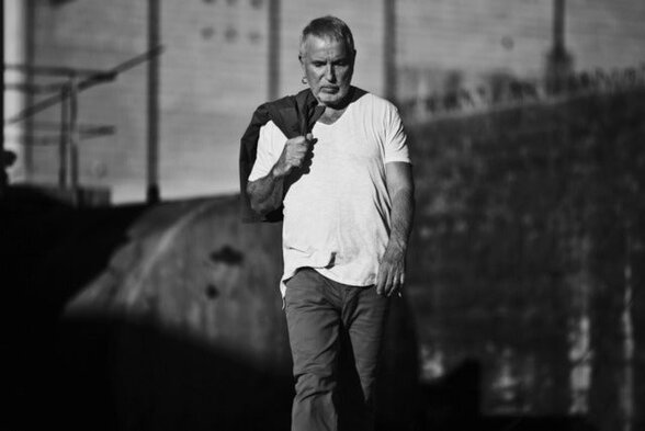 Bernard Lavilliers en noir et blanc, debout tenant une veste sur l'épaule, éclairage de scène en arrière-plan - Agrandir l'image, fenêtre modale