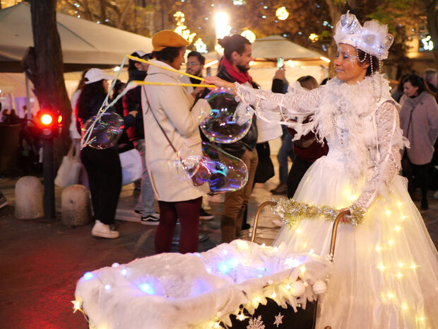Artiste en costume blanc couronné soufflant de grandes bulles de savon depuis un chariot orné de guirlandes lumineuses