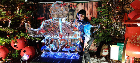 Sculpteur posant devant une sculpture de glace illuminée en bleu représentant le chiffre 2025, dans un décor de marché de Noël - Agrandir l'image, fenêtre modale