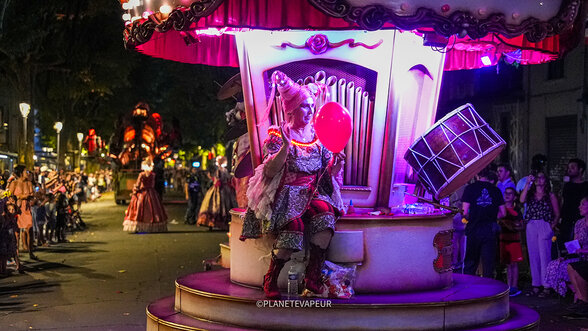 Artiste en costume baroque perchée sur un manège illuminé tenant un ballon rouge, devant la foule en fête - Agrandir l'image, fenêtre modale