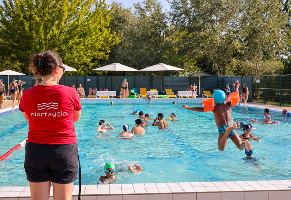 Une maitre nageuse de Niort Agglo encadre les baigneurs dont l'un saute dans le bassin d'une piscine extérieure ensoleillée. - Agrandir l'image, fenêtre modale