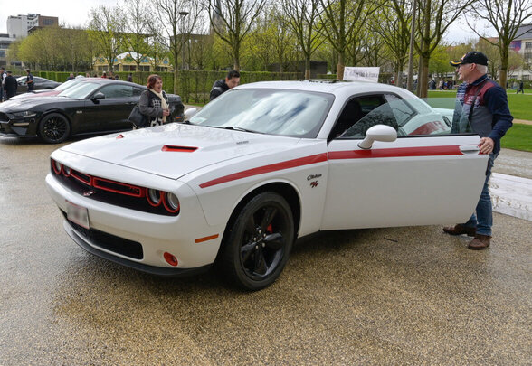 Dodge Challenger blanc à bandes rouges présenté en extérieur, un visiteur à ses côtés, d'autres muscle cars américains visibles en arrière-plan - Agrandir l'image, fenêtre modale