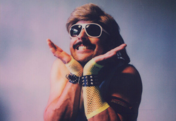 Michel Mercury adopte une pose scénique expressive et souriante, lunettes de soleil blanches et accessoires de scène colorés - Agrandir l'image, fenêtre modale