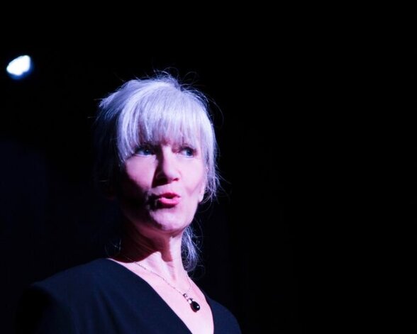 Nina Gomez, conteuse, en pleine narration sur scène, visage expressif éclairé par les projecteurs sur fond sombre - Agrandir l'image, fenêtre modale