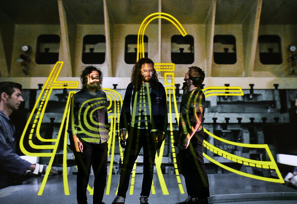 Les trois membres du groupe devant un décor industriel, avec une typographie néon jaune « trafic » projetée sur eux - Agrandir l'image, fenêtre modale