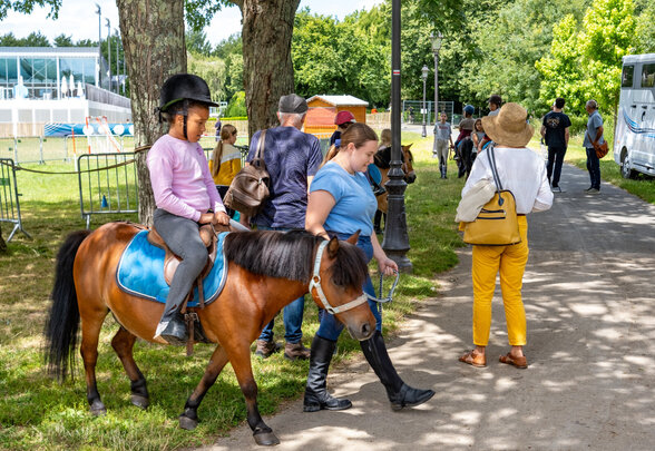 Enfant avec casque équestre faisant une balade à poney, accompagné d'une monitrice, lors de Niort Plage - Agrandir l'image, fenêtre modale