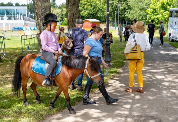 Enfant casqué en selle sur un poney brun, guidé par un animateur en tenue équestre sur un chemin ombragé. - Agrandir l'image, fenêtre modale