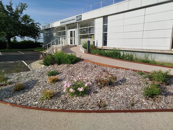 Façade extérieure du bâtiment moderne blanc avec entrée vitrée, massifs fleuris et allée d'accès en premier plan - Agrandir l'image, fenêtre modale