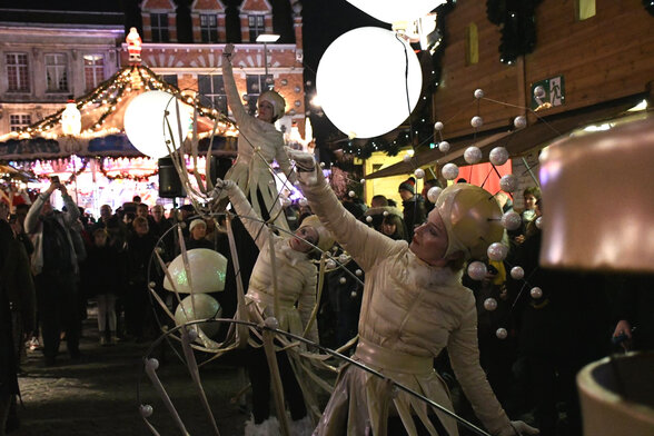 Artistes en costumes blancs manipulant de grandes sphères lumineuses devant un marché de Noël nocturne - Agrandir l'image, fenêtre modale