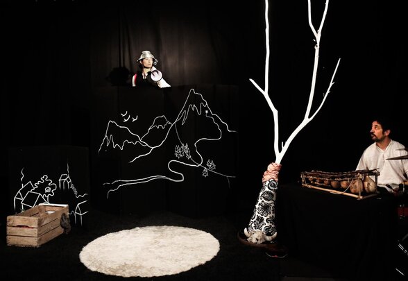 Scène de spectacle : comédienne au casque argenté tenant un mégaphone, musicien aux percussions, arbre lumineux et dessins au trait sur fond noir - Agrandir l'image, fenêtre modale