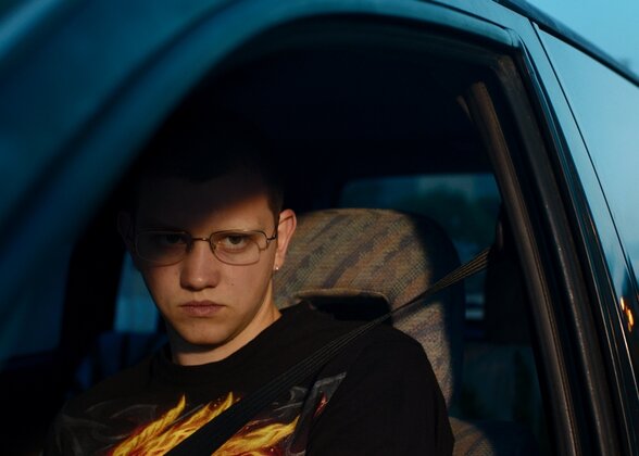 Teddy, jeune homme aux lunettes et t-shirt à flammes, regard sombre et menaçant depuis l'habitacle d'une voiture - Agrandir l'image, fenêtre modale