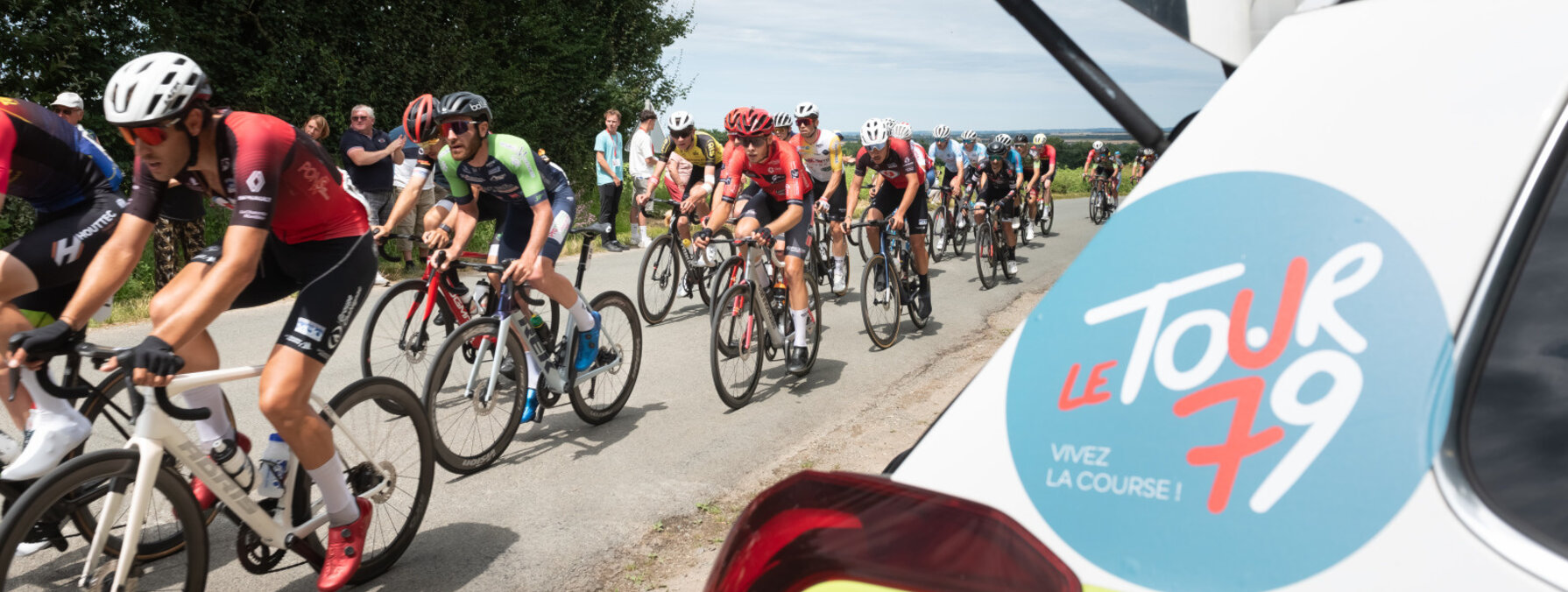 Peloton de cyclistes lancé sur une route de campagne, véhicule officiel Tour 79 au premier plan