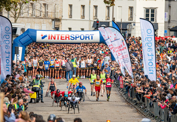 Départ de la course sous l'arche INTERSPORT, athlètes handisport en fauteuil en tête parmi une foule de coureurs et spectateurs - Agrandir l'image, fenêtre modale