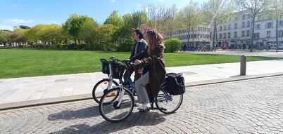 Deux cyclistes, un homme et une femme, circulant côte à côte sur les pavés d'une place en plein soleil printanier