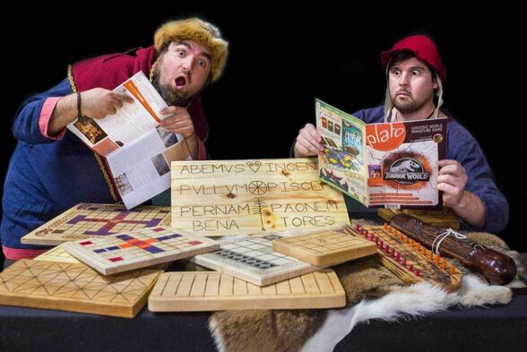 Deux comédiens en tenue médiévale, entourés de jeux anciens en bois gravé et magazines modernes, avec des expressions théâtrales étonnées - Agrandir l'image, fenêtre modale