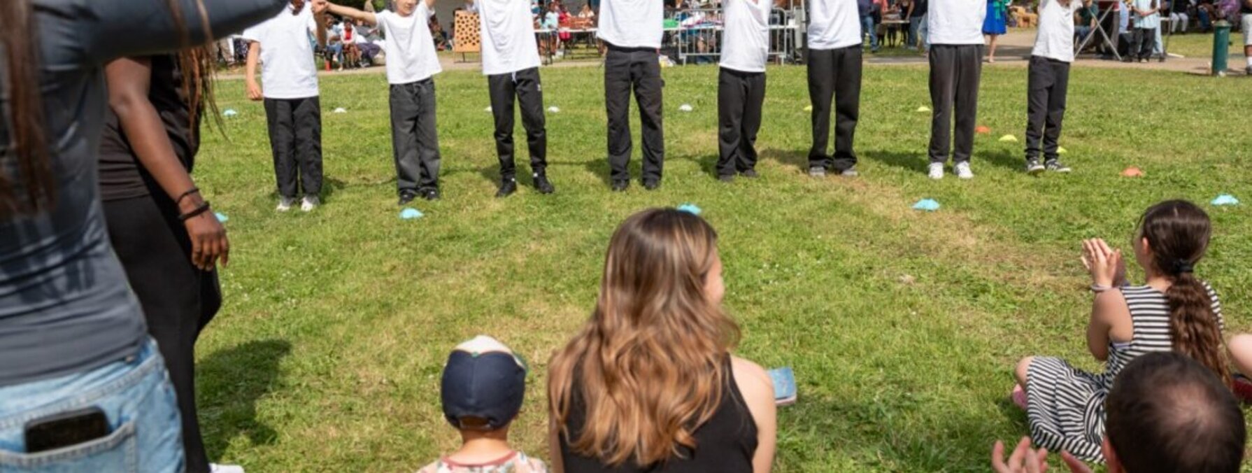 Groupe de jeunes bras levés et mains jointes lors d'une animation associative en plein air, devant un public réuni dans un parc.