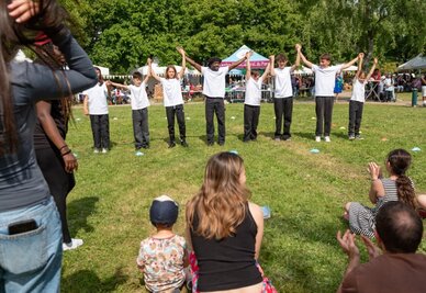 Groupe de jeunes bras levés et mains jointes lors d'une animation associative en plein air, devant un public réuni dans un parc.
