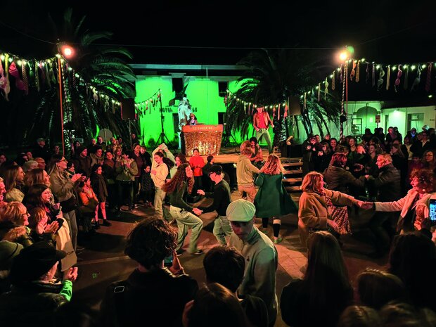 Public formant une ronde festive autour de danseurs en plein air, de nuit, sous des guirlandes lumineuses et des palmiers éclairés en vert.