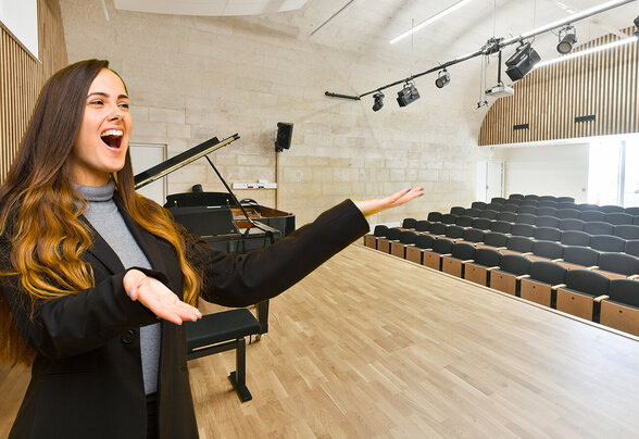 Étudiante chantant sur scène, bras ouverts, dans une salle de concert vide équipée d'un piano à queue et de projecteurs - Agrandir l'image, fenêtre modale