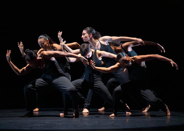 Six danseurs en tenues noires sur scène, corps inclinés en synchronie, bras tendus dans un élan collectif - Agrandir l'image, fenêtre modale