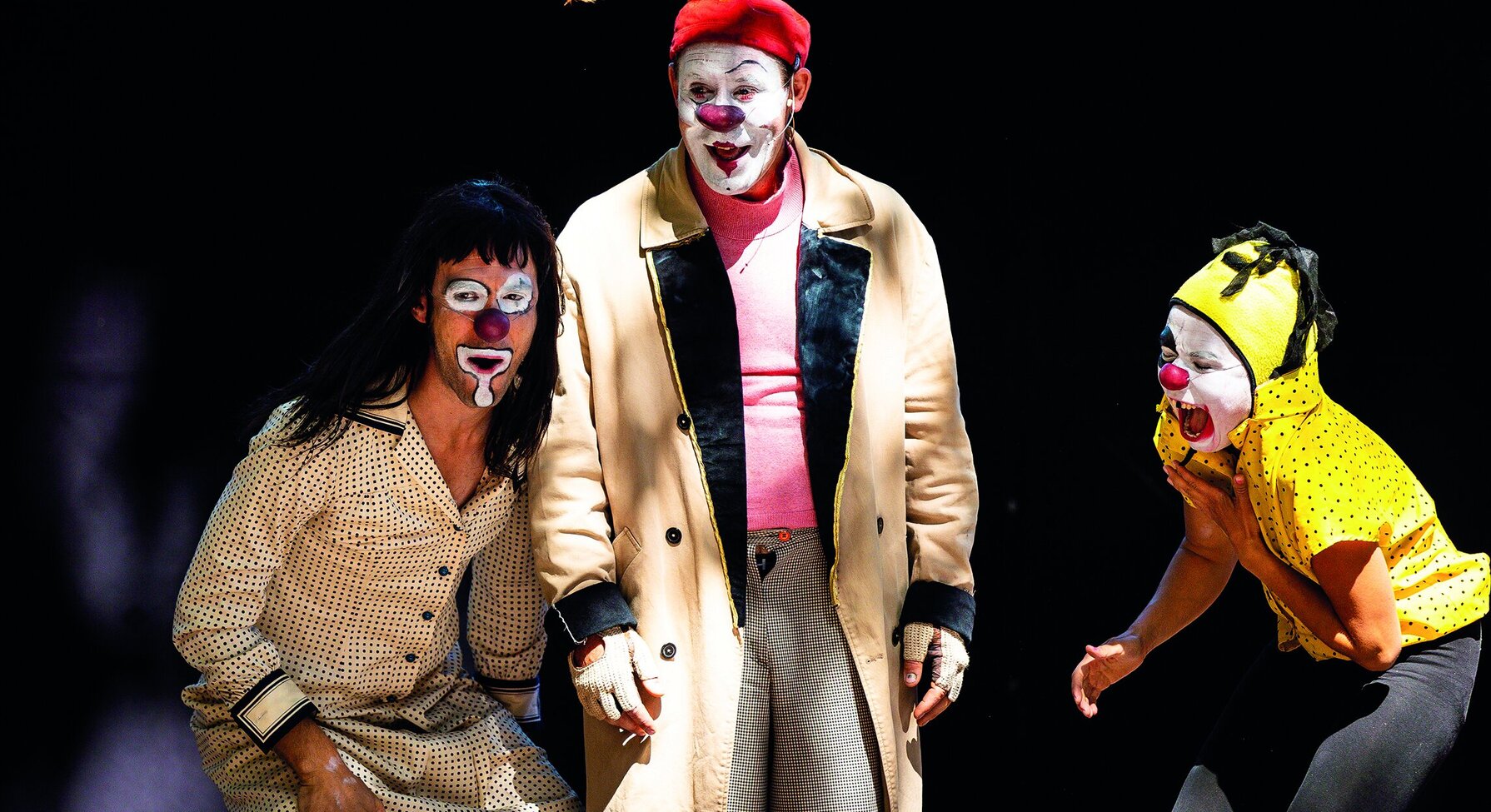 Trois clowns maquillés en costumes colorés jouent une scène comique sur un plateau sombre