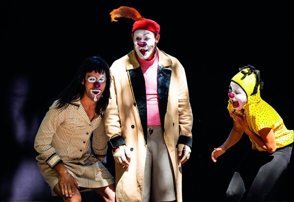 Trois clowns maquillés en costumes colorés jouent une scène comique sur un plateau sombre - Agrandir l'image, fenêtre modale