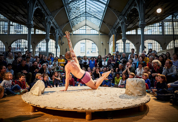 Nathan Israël, danseur et jongleur, en équilibre acrobatique sur une scène ronde couverte d'argile devant un public enthousiaste - Agrandir l'image, fenêtre modale