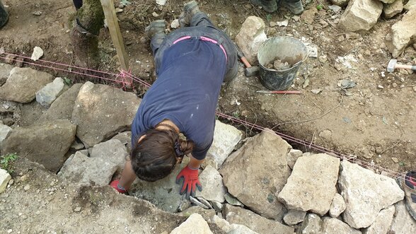 Un jeune participant manipule des pierres avec des gants de protection lors de travaux de maçonnerie en pierres sèches. - Agrandir l'image, fenêtre modale