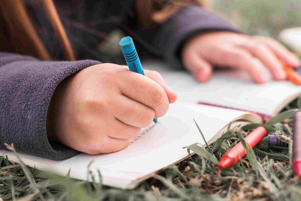 Main d'enfant tenant un crayon bleu pour écrire dans un carnet posé sur l'herbe - Agrandir l'image, fenêtre modale