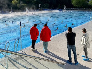 Quatre membres du staff observent depuis le bord les nageuses s'entraînant en couloirs dans le bassin extérieur.