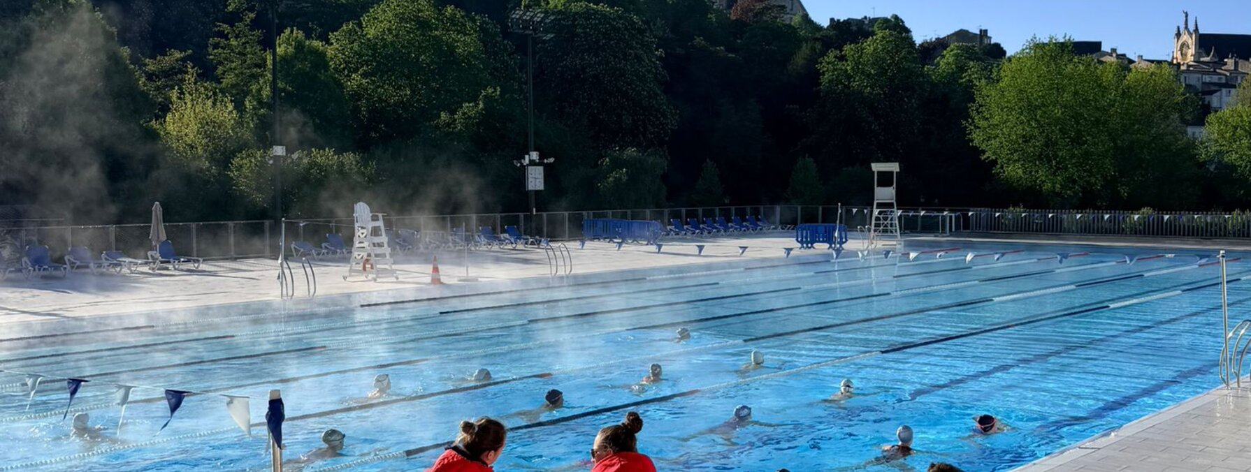 Quatre membres du staff observent depuis le bord les nageuses s'entraînant en couloirs dans le bassin extérieur.