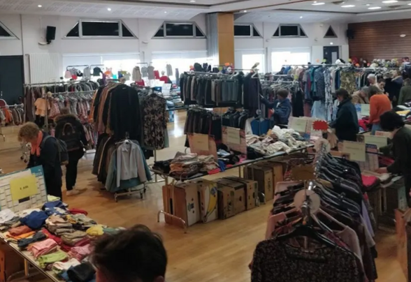 Salle de braderie avec des stands de vêtements sur portants et tables, et des visiteurs en train de chiner