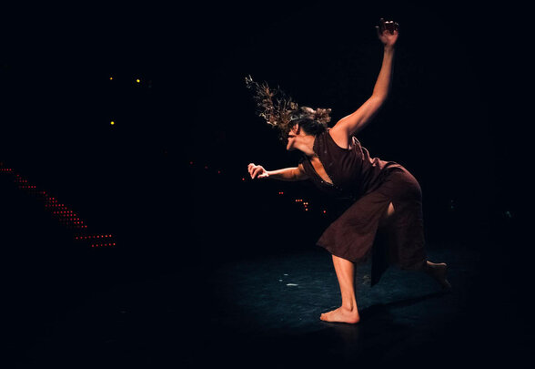 Danseuse en mouvement sur scène, bras levé, coiffure ornée de branchages, dans un éclairage sombre - Agrandir l'image, fenêtre modale