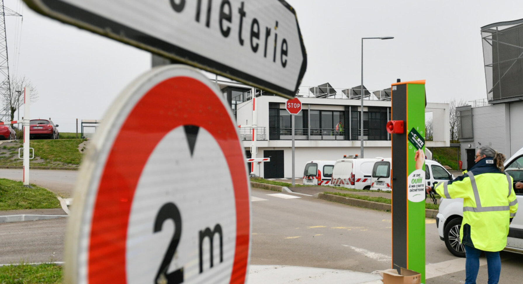 Technicien en gilet fluorescent opérant une borne d'accès verte à l'entrée du centre, un véhicule en attente