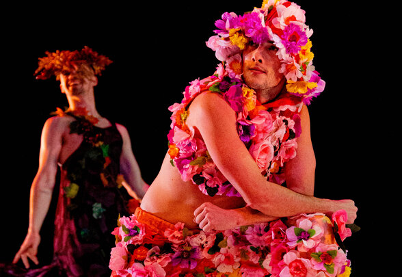 Deux artistes vêtus de costumes floraux multicolores et de couronnes de fleurs, évoluant sur scène dans une lumière chaude - Agrandir l'image, fenêtre modale