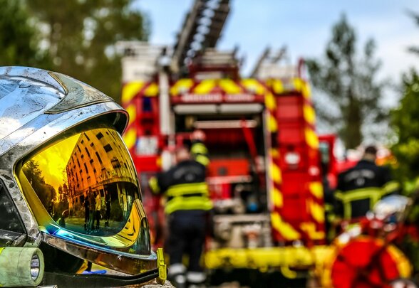 Casque chromé de sapeur-pompier au premier plan, camion grande échelle déployé et pompiers en action à l'arrière-plan - Agrandir l'image, fenêtre modale