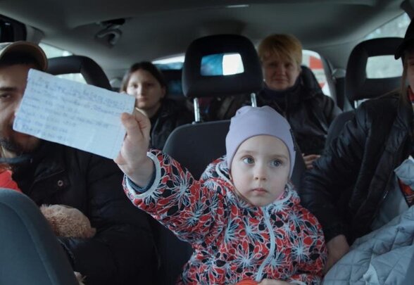 Scène du film : une jeune enfant tend un document manuscrit à la main, entourée de passagers à bord d'un véhicule - Agrandir l'image, fenêtre modale