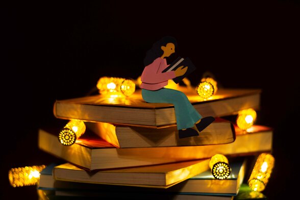 Figurine en papier lisant un livre sur une pile de livres illuminée par des guirlandes lumineuses dorées, ambiance nocturne et chaleureuse - Agrandir l'image, fenêtre modale
