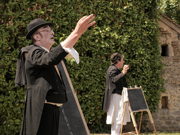 Deux artistes en costume noir et chapeau melon jouent en plein air, l'un main levée, l'autre près d'un tableau noir sur chevalet. - Agrandir l'image, fenêtre modale