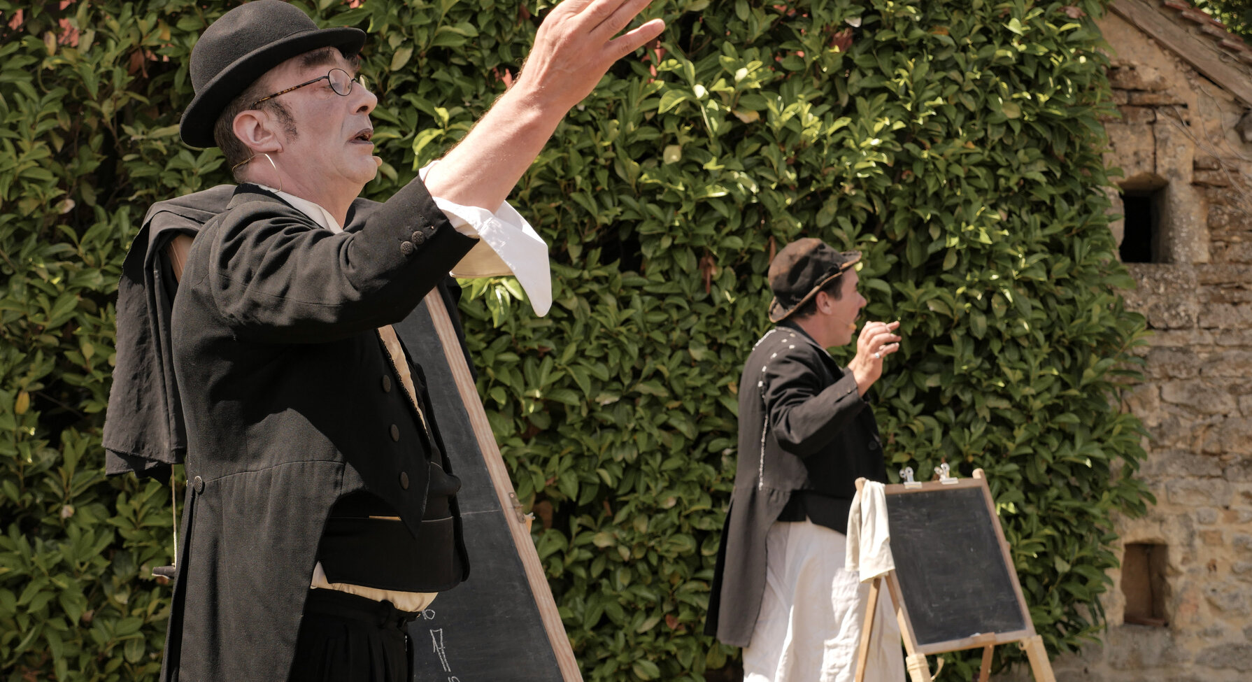 Deux artistes en costume noir et chapeau melon jouent en plein air, l'un main levée, l'autre près d'un tableau noir sur chevalet.