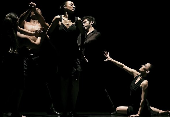 Cinq danseurs sur scène, certains enlacés, une danseuse au sol tendant le bras vers le groupe, dans un éclairage dramatique noir - Agrandir l'image, fenêtre modale