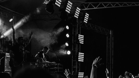 Artistes sur scène sous les projecteurs et la fumée, public les bras levés, ambiance de concert en noir et blanc - Agrandir l'image, fenêtre modale