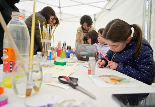 Enfants concentrés sur des créations manuelles, entourés de pinceaux, peintures et ciseaux, encadrés par des adultes sous un chapiteau - Agrandir l'image, fenêtre modale